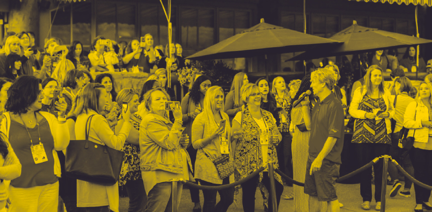 A large group of women gathered outside a building, some taking photos and others listening to a person speaking into a microphone, all under a yellow monochromatic filter.