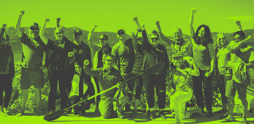 A group of thirteen people with raised fists in a triumphant pose, standing on a dock with a lake and mountains behind them, all in a green monochromatic filter.