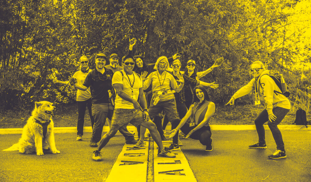 Nine people and a dog pose joyfully on a road marked "FINISH," amidst greenery, all smiling and gesturing playfully in a yellow-toned image.